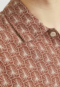 Camisa de manga corta de tela marrón con un patrón abstracto de hojas blancas. Incluye un cierre de botón y un cuello estructurado.