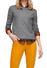 Person, der einen grauen Pullover über einem hochgekrempelten Karohemd und senfgelben Hosen trägt, mit einer Hand in der Tasche, vor einem weißen Hintergrund.