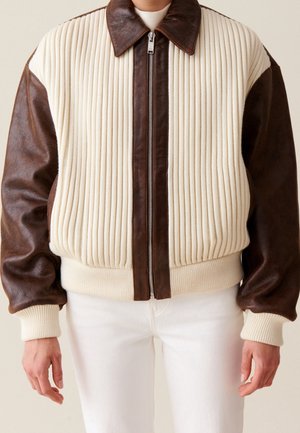 Persoon draagt een crèmekleurig gebreid bomberjack met bruine leren mouwen, kraag en ritssluiting, gecombineerd met een witte broek.