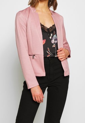 Persoon draagt een lichtroze blazer over een zwart topje met bloemenkant en een zwarte broek, houdt met één hand het revers van de blazer vast.
