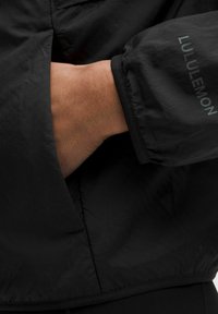 Sorte letvægtsjakke lavet af nylon med elastiske manchetter. Har diskret branding på ærmet og afslappet pasform. Teksturen fremstår glat.