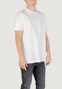 T-shirt bianco in cotone con collo rotondo e maniche corte, dotato di un piccolo patch logo rettangolare sul lato sinistro del petto.