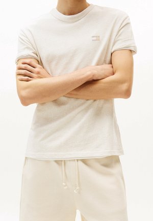 Světlobéžové bavlněné tričko s krátkými rukávy, kulatým výstřihem a malým logem na hrudi. Nošeno s texturovanými béžovými kalhotami.