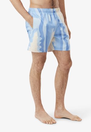 Uomo che indossa pantaloncini con vita elastica con motivo a strisce astratte blu, beige e bianche, in piedi a piedi nudi con le mani nelle tasche.