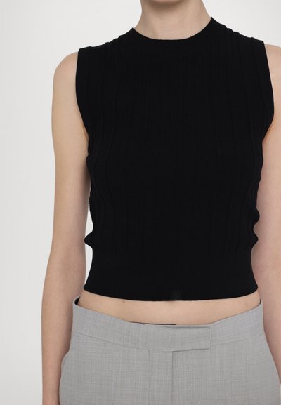 Top noir court sans manches avec une texture côtelée, un col rond et une silhouette ajustée. Porté avec un pantalon gris clair sur mesure.