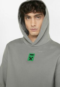 Grå hoodie med en fremtrædende grøn lap, der har et logo og et blokdesign. Blød tekstur, snorhætte og ribbet halsudskæring.