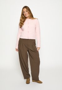 Šviesiai rožinis pūkuotas kardiganas su ilgomis rankovėmis ir apvalia iškirpte, susegamas sagomis priekyje, derinamas su laisvomis rudomis kelnėmis ir rudais batais.