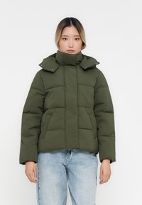 Chaqueta acolchada verde oscuro con un alto cuello, botones a presión y bolsillos laterales. Presenta una forma cuadrada y una textura suave.