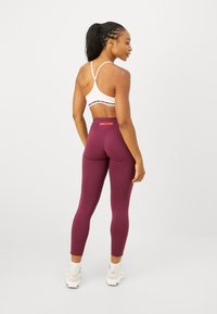 Moteris atsukusi nugarą, vilkinti šviesiai rožinį sportinį liemenėlę, aukštai įsistato maroon atspalvio leginsus, baltas kojines ir smėlio spalvos sportbačius ant balto fono.