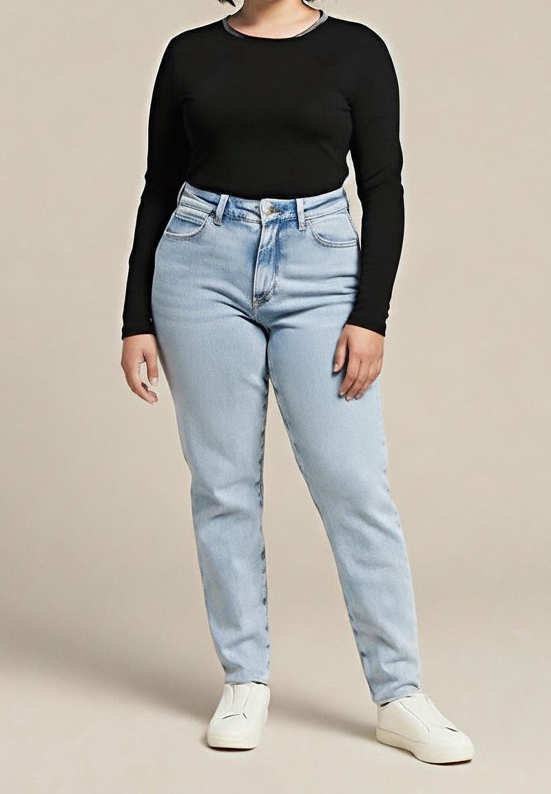 Personne portant un jean taille haute bleu clair, un haut noir à manches longues et des chaussures blanches à enfiler, debout les mains détendues le long du corps.