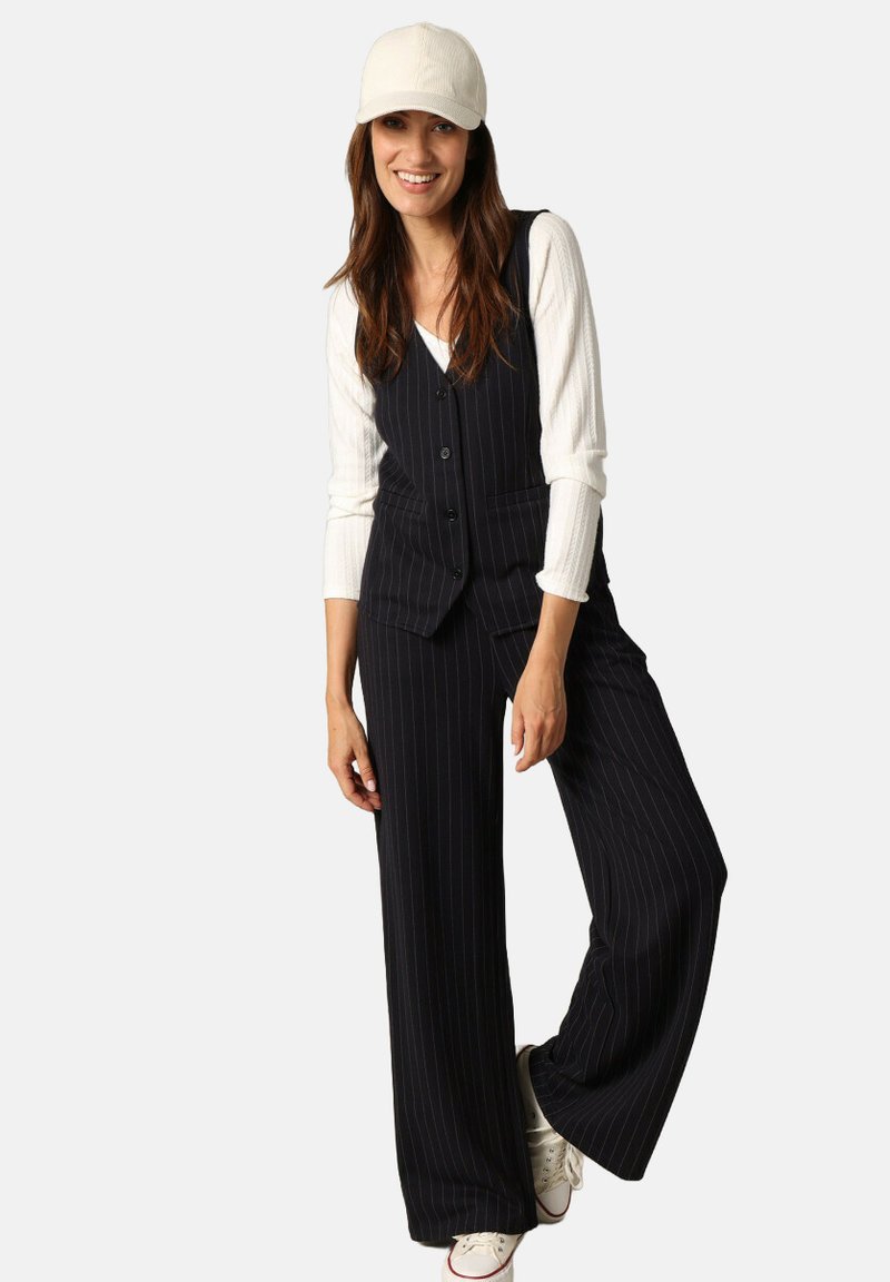 Un gilet à rayures noires et un pantalon large, associé à une chemise à manches longues côtelée blanche et une casquette beige. Des baskets blanches complètent le look.