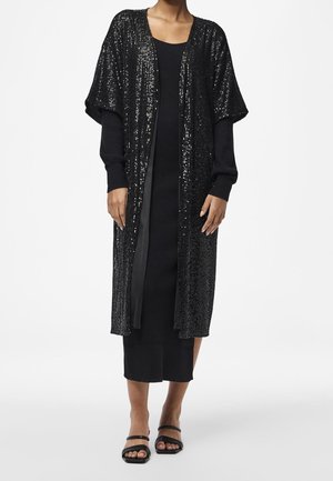 Femme portant un long cardigan noir à sequins sur une robe noire côtelée à manches longues et des sandales noires, debout sur un fond blanc.