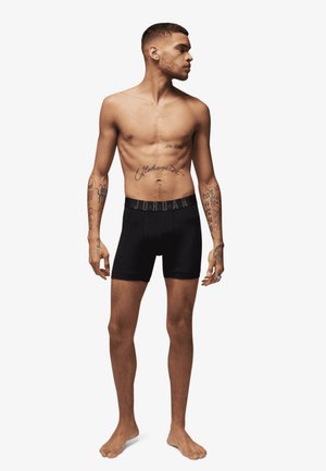 Sorte boxer-shorts med en mærkevare elastik, modellen står barfodet. Synlige tatoveringer på overkroppen og armene, kort hår, neutral ansigtsudtryk.