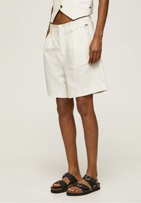 White linen shorts with a pleated design, side pockets, and a relaxed fit, paired with black sandals featuring straps and circular buckles.