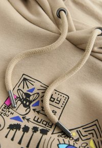 Beige hættejakke med en struktureret stof, der har en snor med metalstop og farverige grafiske prints af mønstre og symboler på forsiden.