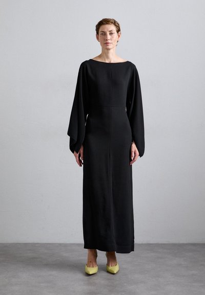 Lang, zwart jurk met een bootneklijn en oversized mouwen, gemaakt van soepel stof. Gecombineerd met gele puntige hakken.
