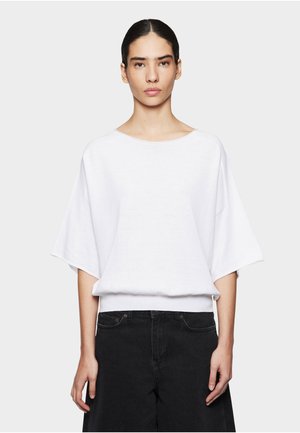 Vrouw met donker haar in een staart draagt een los wit shirt met korte mouwen en zwarte hoge taillebroek, staand tegen een effen lichte achtergrond.