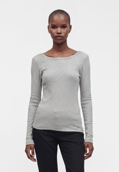 Femme aux cheveux très courts portant un haut gris clair à manches longues, près du corps, et un pantalon noir, debout devant un fond blanc uni.
