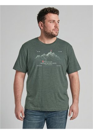 Homme aux cheveux courts et bouclés portant un t-shirt graphique vert avec un motif de montagne et le texte "Explore Beyond Limits" ainsi qu'un jean bleu.