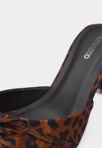 Mule à talons hauts avec imprimé léopard marron et noir, semelle intérieure noire, et un petit nœud décoratif sur le devant.