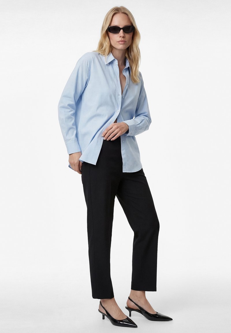 Blonde Frau mit schwarzen Sonnenbrillen, hellblauem, geknöpftem Hemd, schwarzen, kurzen Hosen und schwarzen Slingback-Schuhen mit kleinem Absatz, stehend.