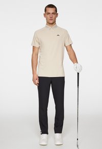 Beige pikétröja, svart logotyp, kort ärm. Svarta byxor, slät textur. Vita golfskor. Modellen håller en golfklubba med vit handske.