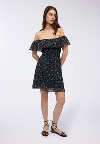 Abito nero con spalle scoperte, fantasia floreale blu, maniche a volant trasparenti, corpetto arricciato e gonna svasata. Abbinato a sandali neri con fasce.