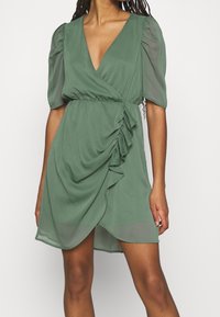 Robe enveloppante verte avec un décolleté en V profond, des manches courtes bouffantes et un détail de volants sur le côté. Tissu léger, longueur au genou, texture semi-transparente.