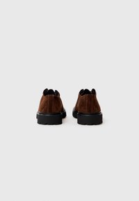 Une paire de chaussures en suède marron avec des semelles en caoutchouc noir épais, présentée de l'arrière sur un fond clair.