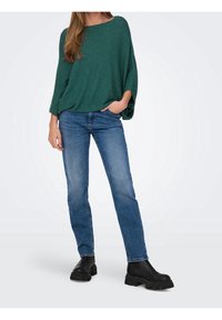 Haut en maille vert, ample, à manches larges, associé à un jean bleu à jambes droites et des bottines noires à semelle épaisse.