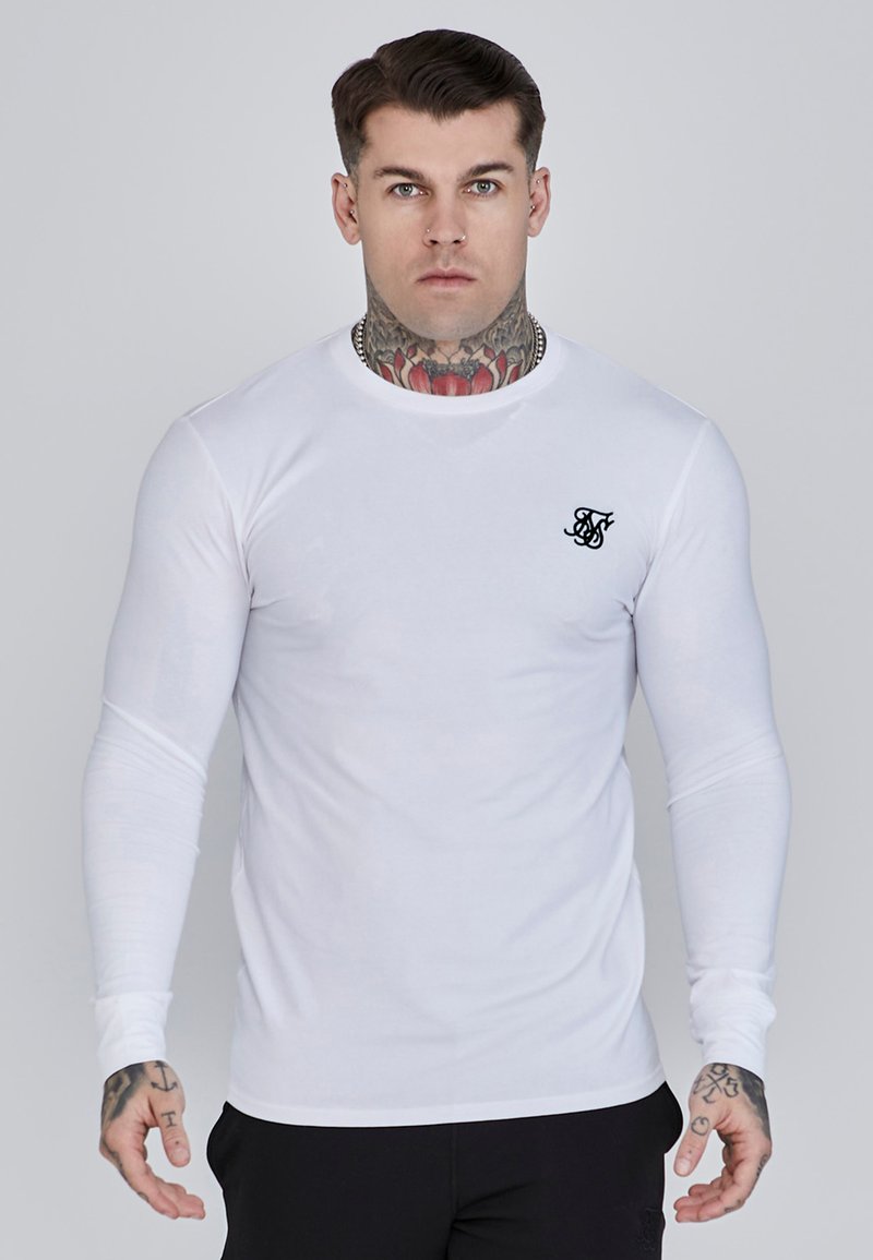 Homme avec tatouages sur le cou et la main, portant une chemise blanche à manches longues avec un logo noir, debout devant un fond gris uni.
