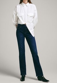 Camicia bianca a maniche lunghe con due tasche sul petto, abbinata a jeans blu scuro a gamba dritta. Stivaletti neri con punta a contrasto completano l'outfit.