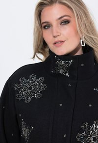 Femme blonde portant un manteau noir orné de motifs de flocons de neige en sequins et perles complexes, accompagné de boucles d'oreilles rectangulaires en argent.