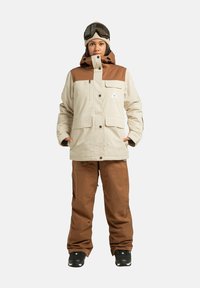 Kurtka narciarska w kolorze kremowym z brązowymi akcentami, dwie kieszenie na piersi oraz zamek błyskawiczny. Do tego brązowe spodnie narciarskie i czarne buty z fakturowaną powierzchnią.