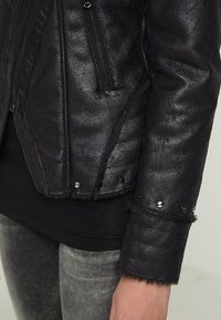 Veste noire en similicuir avec un design côtelé, poches zippées et clous argentés le long de l'ourlet. Surface texturée avec une bordure duveteuse aux poignets.