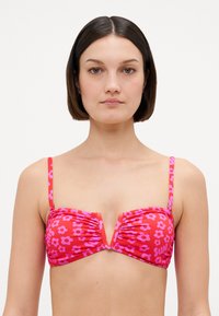 Mulher com cabelo curto e escuro, vestindo um top de biquíni vermelho com padrão floral rosa e alças finas, de frente contra um fundo branco.