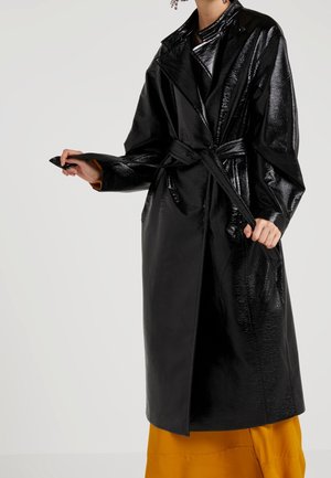 Sort, blankpoleret trenchcoat med belted talje, brede reverser og sidelommer, båret over en sennepsgul nederdel. Glat, blank tekstur.