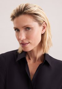 Blondynka o krótkich włosach, ubrana w czarną koszulę z kołnierzykiem, patrząca lekko w bok na jasnym, jednolitym tle.