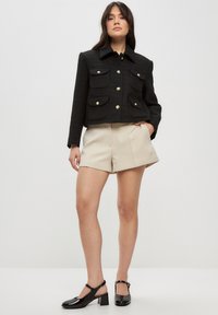 Chaqueta negra corta con cuatro bolsillos grandes y botones plateados, combinada con pantalones cortos beiges y zapatos de tacón negro brillantes.