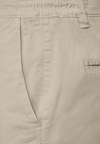 Gros plan sur un pantalon beige montrant la poche avant, la passante de ceinture et les coutures détaillées sur le tissu.