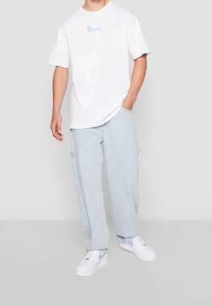 Homme portant un jean bleu clair ample, un t-shirt blanc oversize avec un petit logo bleu, et des baskets blanches avec des touches bleues, debout avec une main dans la poche.