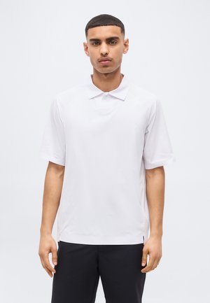 Noor mees lühikeste juuste ja tavalise valge lühikese varrukaga polosärgiga ning mustade pükstega, seismas heleda tausta ees.