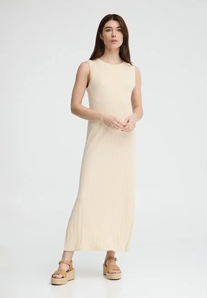 Kobieta stojąca w kremowej, bezrękawowej sukni maxi i beżowych sandałach na platformie z paskami wokół kostek na białym tle.