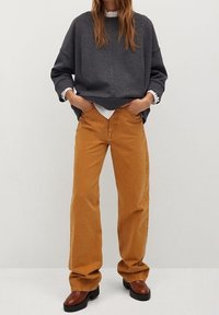 Sweatshirt gris surdimensionné, chemise blanche en dessous, pantalon orange à jambes larges, chaussures brunes, pose debout avec les mains dans les poches.