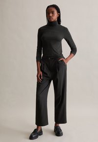 Schwarzes, tailliertes Rollkragenshirt mit glatter Textur, kombiniert mit weiten schwarzen Hosen. Das Model trägt klobige schwarze Loafers. Neutraler Hintergrund.