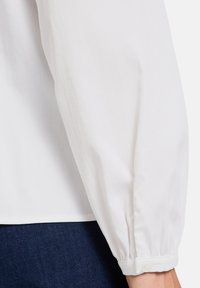 Hvid bluse med en samlet, puffet ærme. Har en manchet med en knaplukning. Glat, letvægtsstof.