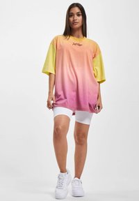 Overdimensjonert t-skjorte i gradientfarger fra gul til rosa, med kortermet og mykt stoff, kombinert med hvite sykkelshorts og joggesko.