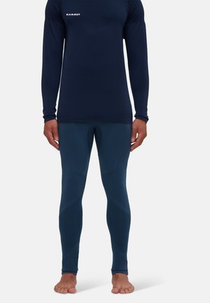 Blå Mammut långärmad topp och matchande leggings som bärs av en person som står barfota mot en enkel vit bakgrund.