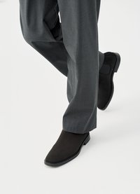 Μαύρα μοκασίνια από σουέντ με λείο φινίρισμα, με στρογγυλερή μύτη και επίπεδη σόλα από καουτσούκ, συνδυασμένα με γκρι παντελόνια ραμμένα κατά παραγγελία.