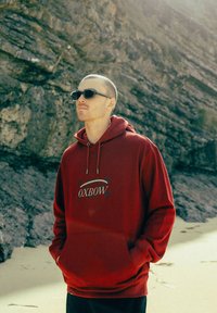 Un sweat à capuche rouge avec une poche frontale et un cordon de serrage ; arbore le logo "OXBOW" en bleu. Porté avec des lunettes de soleil noires sur fond rocheux.
